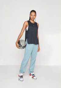 Schwarzes, ärmelloses Sporttop und hellblaue Jogginghose, hält einen Basketball, dazu weiße und rote Sneakers. Kurze Seitenschlitze im Top.