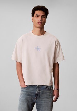 Jeune homme aux cheveux bouclés portant un t-shirt beige Calvin Klein et un jean bleu, debout devant un fond gris uni avec une main dans la poche.