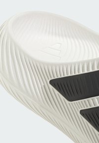 Zbliżenie na białą, teksturowaną podeszwę buta z czarnymi gumowymi uchwytami i subtelnym tłoczonym trójkątnym logo w obszarze pięty.