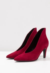 Zapatos de tacón alto de ante rojo con punta afilada, diseño de corte profundo en V y un tacón negro delgado, con una textura suave.