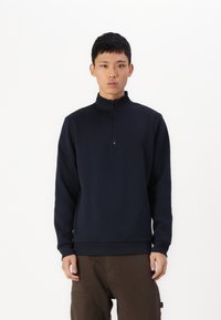 Pullover navy blu con zip realizzato in un tessuto morbido; presenta un colletto alto e polsini a coste, abbinato a pantaloni marroni.