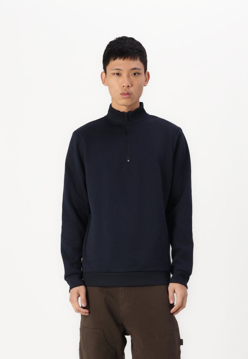 Pullover navy blu con zip realizzato in un tessuto morbido; presenta un colletto alto e polsini a coste, abbinato a pantaloni marroni.