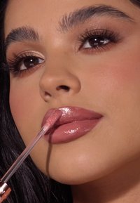 Błyszczyk do ust o błyszczącym, jasnoróżowym odcieniu nałożony aplikatorem. Modelka ma gładką skórę i wyraźnie zarysowane brwi, z delikatnym makijażem.