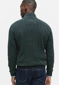 Mörkgrön kabelstickad tröja med hög krage och ribbade ärmslut, med ett strukturerat mönster längs ryggen.