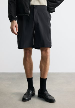 Férfi fekete cipzáras rövidnadrágban, fekete zokniban, fekete mokaszinban és fekete dzsekiben, egyszínű világos háttér előtt.