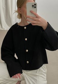 Črno skrajšano jakno z okroglim izrezom, ki ima velike zlate cvetlične gumbe, široke rokave in gladko teksturo. Oblečeno je nad belimi hlačami.