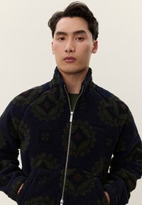 Μπλε-ναυτικό φλις μπουφάν με φερμουάρ μπροστά, που διαθέτει γεωμετρικό σχέδιο σε πράσινο και καφέ. Έχει ψηλό γιακά και δύο μπροστινές τσέπες.