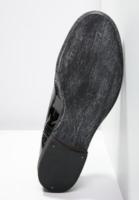 Chaussure en cuir verni noir vue de dessous, présentant une semelle texturée avec de légers motifs et une finition lisse sur la partie supérieure.