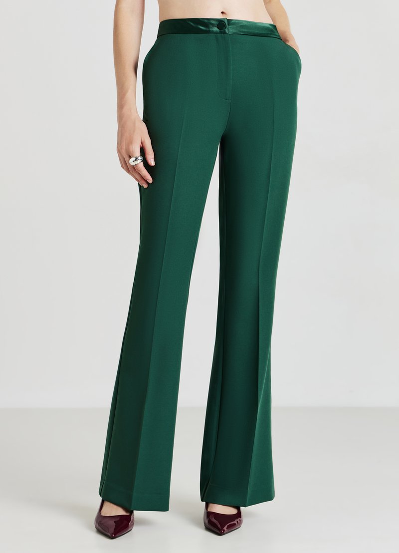 Pantaloni larghi verdi e fluido con una cintura in satin liscia, dotati di chiusura frontale con zip e tasche laterali. Colore uniforme, senza fantasie.