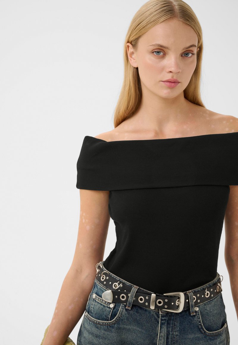 Femme blonde portant un haut noir à épaules dénudées, un jean bleu et une ceinture noire avec des clous en argent et des décorations de notes de musique.