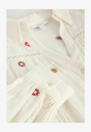 Cremefarbene Bluse mit einem Knopfkragen, die mit roten und braunen gestickten Quadraten sowie plissierten Ärmeln verziert ist. Weicher Stofftextur.