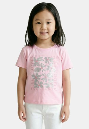 Jeune fille aux cheveux noirs et raides, portant un T-shirt rose avec des motifs floraux argentés et un pantalon blanc, debout devant un fond uni.