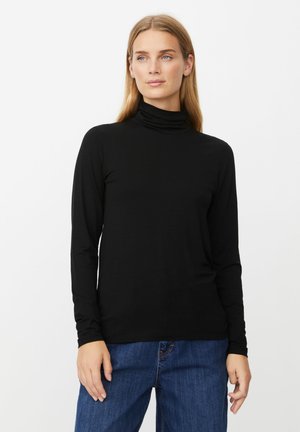 Mujer vestida con un top negro de manga larga y cuello alto, y jeans azules, de pie contra un fondo blanco liso, mirando ligeramente hacia la derecha.