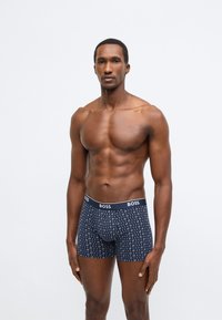 Tengerészkék, pamut boxeralsó, geometriai mintával és márkás derékpánttal. Testhez simuló szabású, combközépig érő fazon.