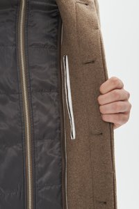 Manteau en mélange de laine marron avec une doublure lisse et sombre. Il est doté d'une fermeture éclair métallique, d'une poche intérieure avec un galon blanc, et de coutures régulières en guise d'accents.