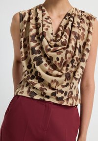 Mouwloos topje met bruine en zwarte luipaardprint en gedrapeerde halslijn, gedragen met high-waisted bordeauxrode broek.