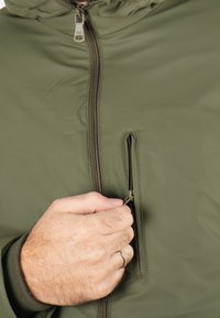 Veste vert olive avec une fermeture éclair frontale, une petite poche zippée et des poignets côtelés. Le tissu semble lisse avec une finition mate.