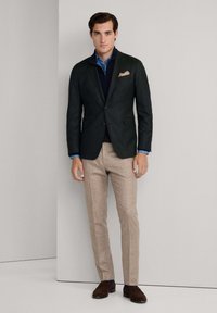 Tamnozelena jakna preko mornarsko plavog pulovera, u kombinaciji s bež krojenim hlačama. Smeđe cipele s plavim detaljima. Maramica u džepu dodaje detalj.