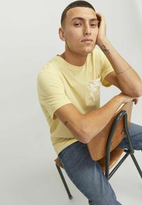 Gul bomulds t-shirt med en mønstret lomme, parret med blå denim jeans. Modellen sidder på en træstol med en neutral baggrund.
