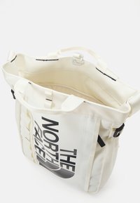 Sac fourre-tout blanc arborant un grand logo noir "The North Face", en tissu résistant, avec plusieurs poignées et des mécanismes de fermeture sécurisés.