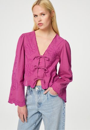 Roze, geborduurde blouse met een diepe V-hals, lange klokmouwen en decoratieve strikjes aan de voorkant, gecombineerd met lichtblauwe denim jeans.