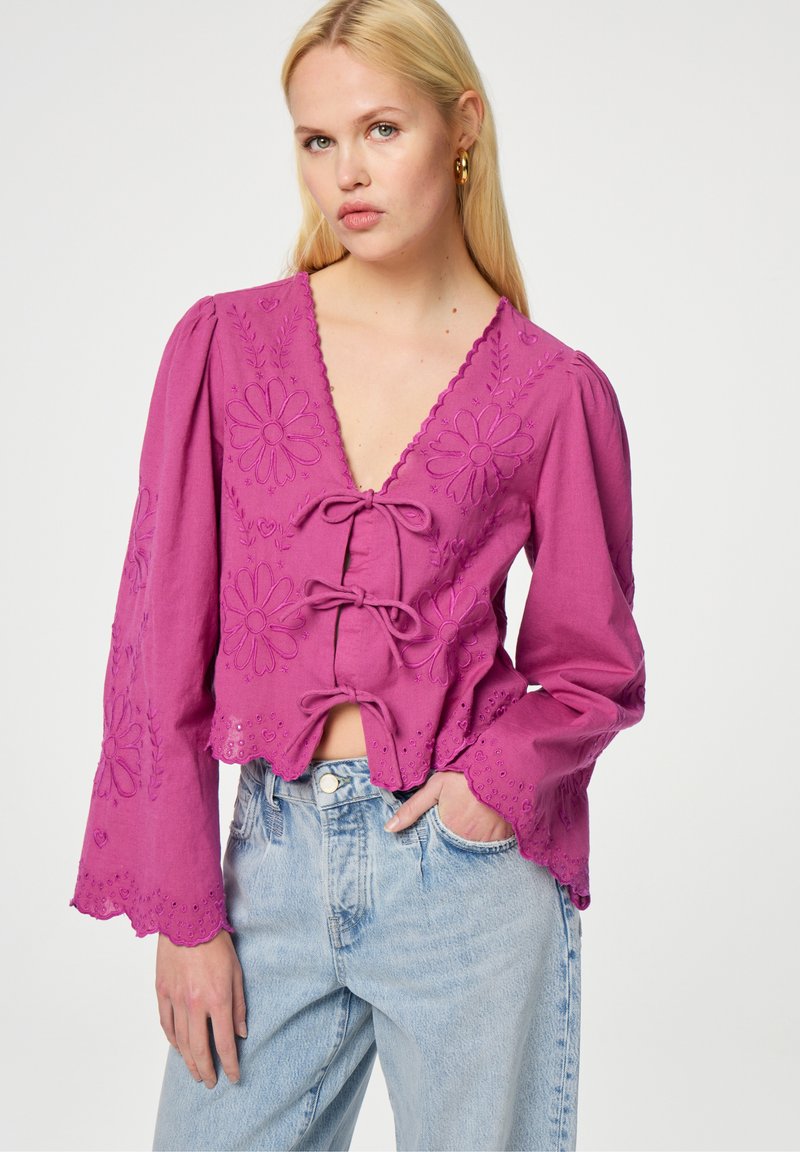 Roze, geborduurde blouse met een diepe V-hals, lange klokmouwen en decoratieve strikjes aan de voorkant, gecombineerd met lichtblauwe denim jeans.