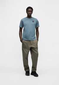 Μπλε βαμβακερό t-shirt με μαύρο λογότυπο, συνδυασμένο με φαρδιά χακί παντελόνια και μαύρα αθλητικά παπούτσια. Απλή, καθημερινή εμφάνιση.