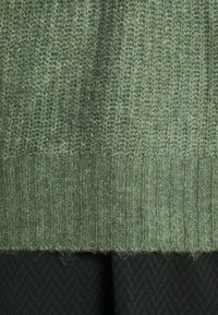 Gros plan sur l'ourlet d’un pull vert en maille sur un pantalon sombre avec un motif chevron subtil.