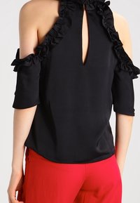 Blusa nera con maniche a fredda spalla a balze, dettaglio a chiave sul retro e tessuto morbido. Abbinata a pantaloni rossi vivaci.