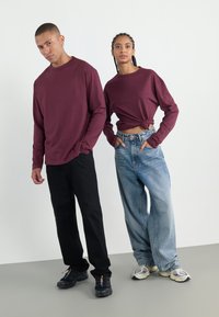 To modeller har på seg burgunderfargede langermede topper. Den ene har på seg løse lyseblåjeans, mens den andre har svarte bukser. Begge har på seg sneakers med varierte design.