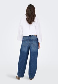 Person with long brown hair wearing a white oversized shirt, loose blue jeans, and brown shoes, standing facing away on a plain background.