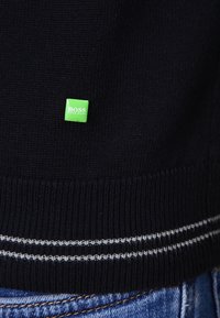 Pull en tricot noir avec un patch logo rectangulaire vert indiquant "BOSS" et des rayures blanches à l'ourlet. La texture est lisse. Du denim est visible en dessous.