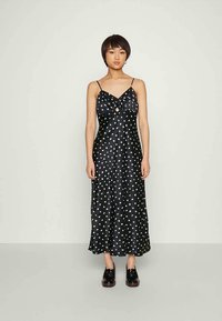 Vestido de cetim preto com bolinhas brancas, apresentando um detalhe frontal torcido e alças finas ajustáveis. Usado com sapatos pretos.