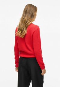 Pull en tricot rouge avec un motif texturé, col rond et manches longues. Porté avec un pantalon noir, vu de dos.