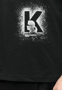 Μαύρο πουκάμισο με μεγάλο λευκό γράμμα "K" με στίγματα και λευκή σιλουέτα ατόμου με αλογοουρά που φοράει γυαλιά ηλίου μέσα στο γράμμα, κείμενο "KARL LAGERFELD JEANS".