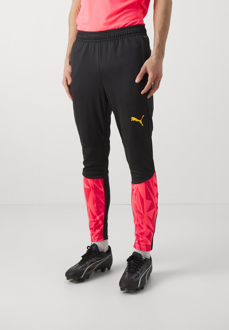 Zwarte sportbroek met een roze geometrisch patroon op de onderbenen en een gele Puma-logo. Gemaakt van lichtgewicht materiaal met een elastische tailleband.