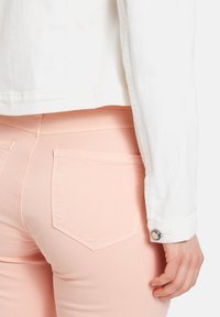 Witte denimjas deels zichtbaar over zachtroze aangesloten jeans met achterzakken. De denimstof lijkt glad met kleine stiksels.