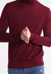 Maglione a dolcevita bordeaux con maniche lunghe, colletto e orlo a coste, realizzato in tessuto morbido, indossato con le mani giunte davanti.