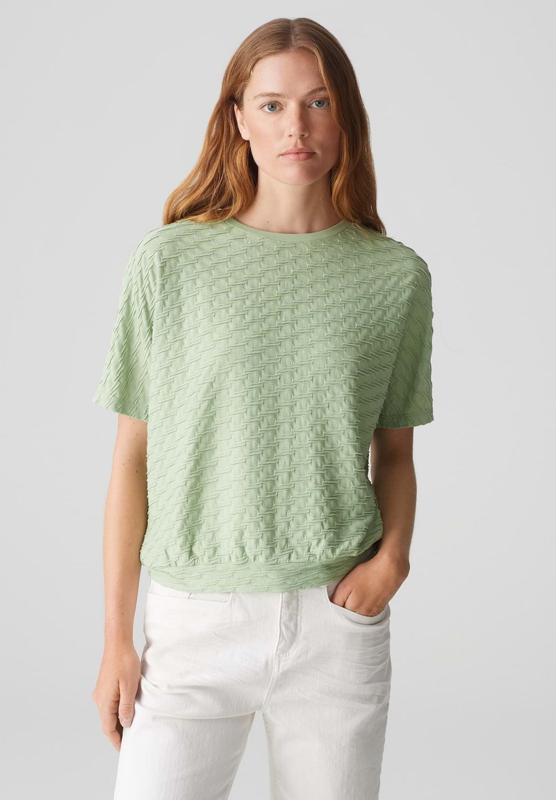 Sweat-shirt vert clair avec un motif texturé, manches courtes, col ras du cou, coupe décontractée, porté avec un pantalon blanc. Design minimaliste.