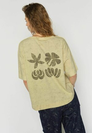 Mujer con cabello ondulado lleva una camisa suelta de color amarillo pálido con cuatro diseños florales abstractos oscuros en la parte posterior y pantalones con patrón oscuro.