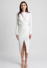 Biała sukienka z długim rękawem, z wycięciem w kształcie dziurki, drapowaną konstrukcją i rozporkiem na wysokości uda; wykonana z gładkiego materiału. Modelka nosi beżowe szpilki.