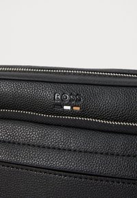 Black textured leather bag close-up showing silver zipper and embossed "BOSS" logo with black, white, and brown rectangular accents.