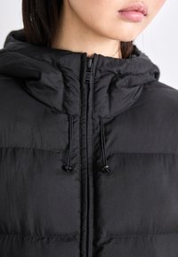 Melnā pildītā jaka ar rāvējslēdzēju, pulksteni ar aukliņu un gludu tekstūru ar šūtām šuvēm siltumam un komfortam.