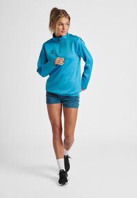 Lichtblauw pullover met een halve rits hals, gecombineerd met teal sportshorts. Heeft een logo op de borst en een ontspannen pasvorm. Zwarte sneakers maken de look af.