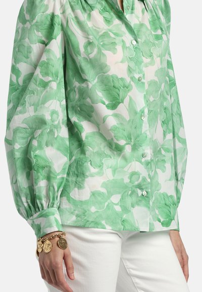 Zaļš un balts ziedu raksta garroku blūze ar pogām un uzpampušām piedurknēm, kombinēta ar baltiem biksēm un zelta piekariņu aproci kreisajā plaukstas locītavā.