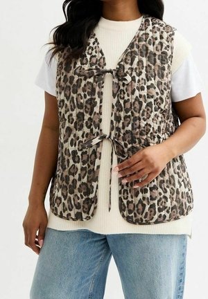 Femme portant une veste sans manches matelassée imprimé léopard nouée devant par deux nœuds, sur un haut blanc côtelé à manches courtes et un jean bleu clair.