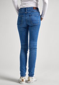 Jeans de mezclilla azul ajustados con un tiro medio, bolsillos traseros y detalles de costura clara. Combinados con zapatillas blancas y doradas.