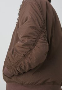 Osoba oblečená v hnědé péřové bundě se shromážděním švu podél rukávu a ribbovanými manžetami, stojící na jednobarevném pozadí.