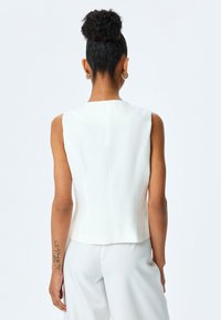 Weißes ärmelloses Oberteil mit hohem Kragen, glatter Textur, geradem Schnitt und Nahtdetails auf der Rückseite. Einfaches und minimalistisches Design.
