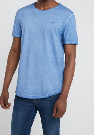Uomo che indossa una T-shirt azzurra a maniche corte con un piccolo logo sul petto e jeans blu scuro, in piedi su uno sfondo semplice.